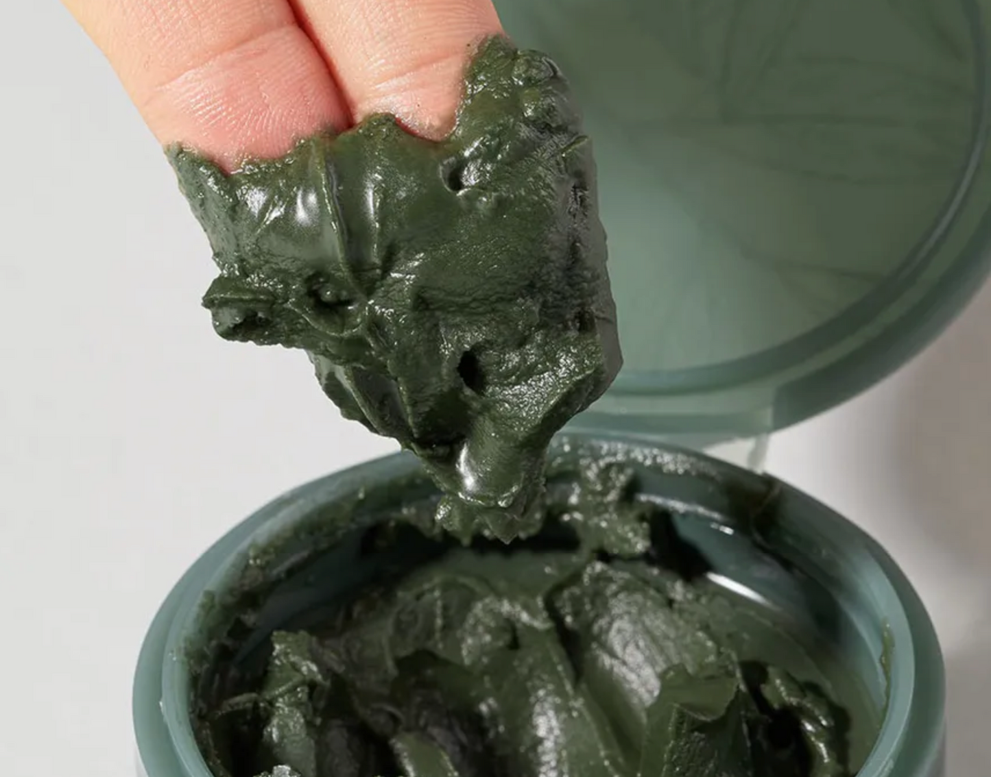 Hand holding a clump of dark green clay above an open container of the same clay.