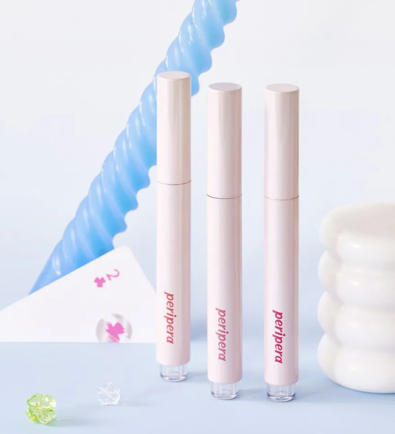 Three Peripera lip glosses on a light blue background with decorative elements.