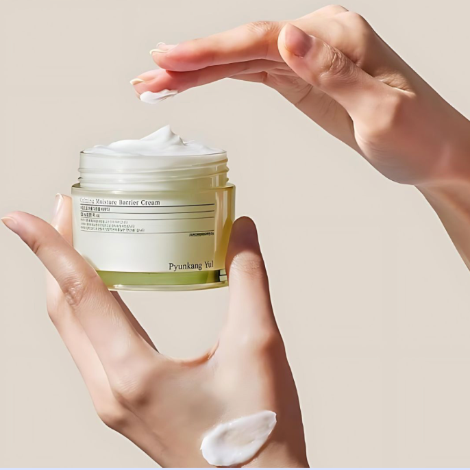 Hand holding a jar of Pyunkang Yul cream with a visible cream smear on a beige background