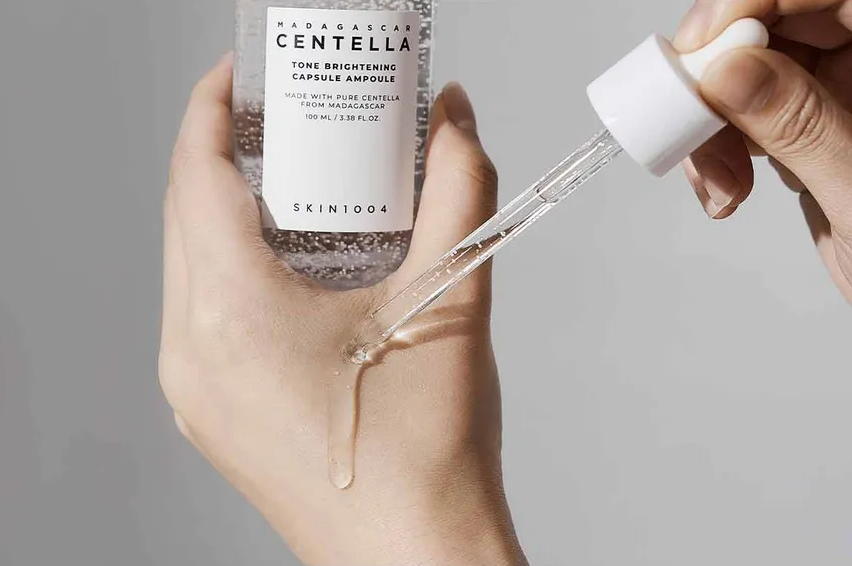 Hand holding a skincare bottle with a dropper, featuring SKIN1004 branding.